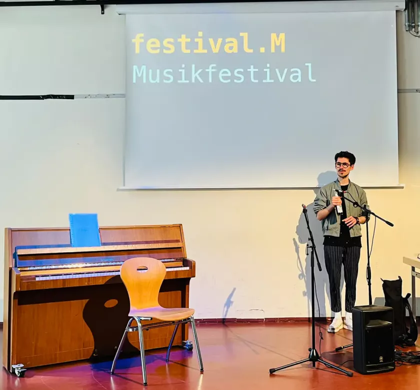 Ein junger Mann spricht beim Musikfestival „festival.M“ auf einer Bühne mit Mikrofon, neben ihm ein Klavier und ein Stuhl.