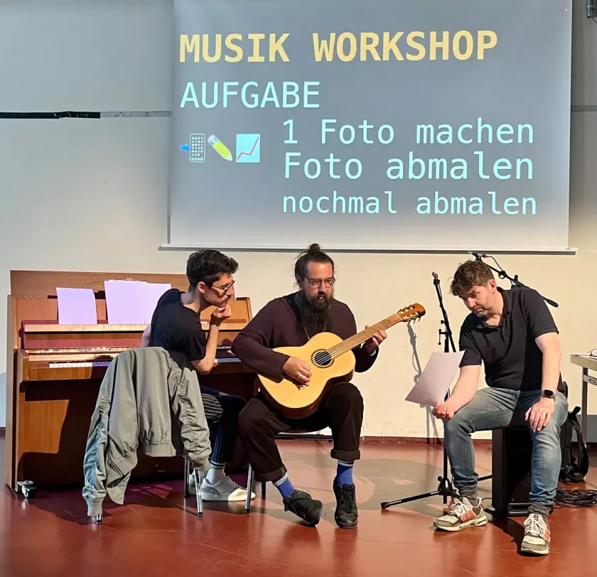 Drei Männer sitzen vor einem Klavier; einer spielt Gitarre, die anderen hören zu, im Hintergrund steht „Musik Workshop“ auf der Leinwand.