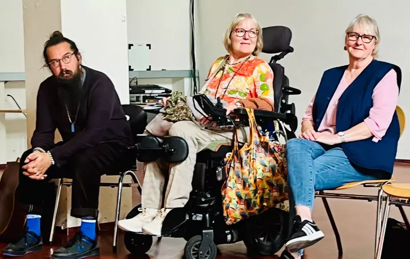 Drei Personen sitzen nebeneinander, darunter eine Frau im Rollstuhl, alle aufmerksam im Workshopgeschehen.